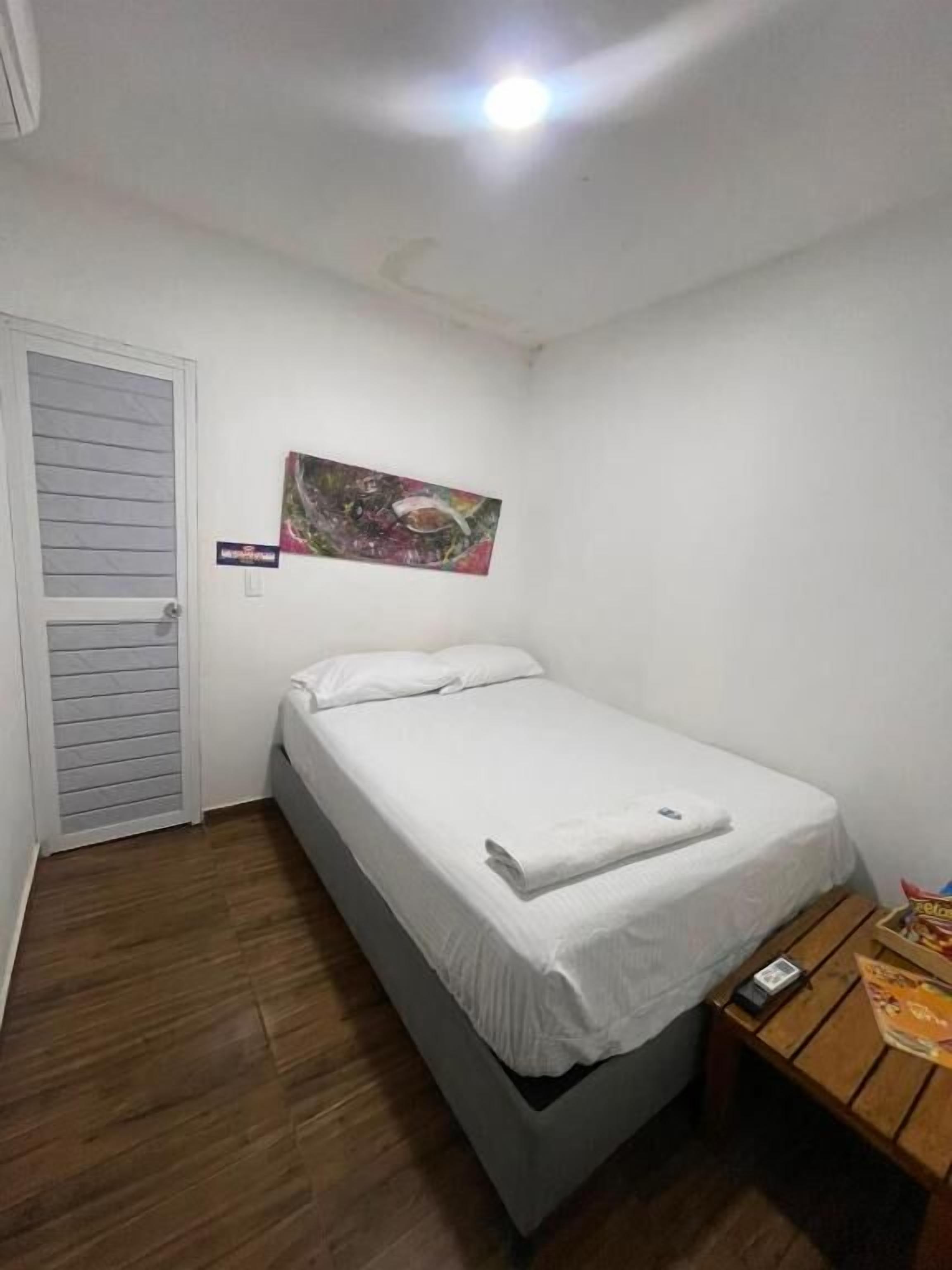 A single-bed room with a wooden floor, a white bed, a small wooden table, and a wall-mounted painting.