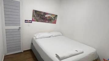 A single-bed room with a wooden floor, a white bed, a small wooden table, and a wall-mounted painting.