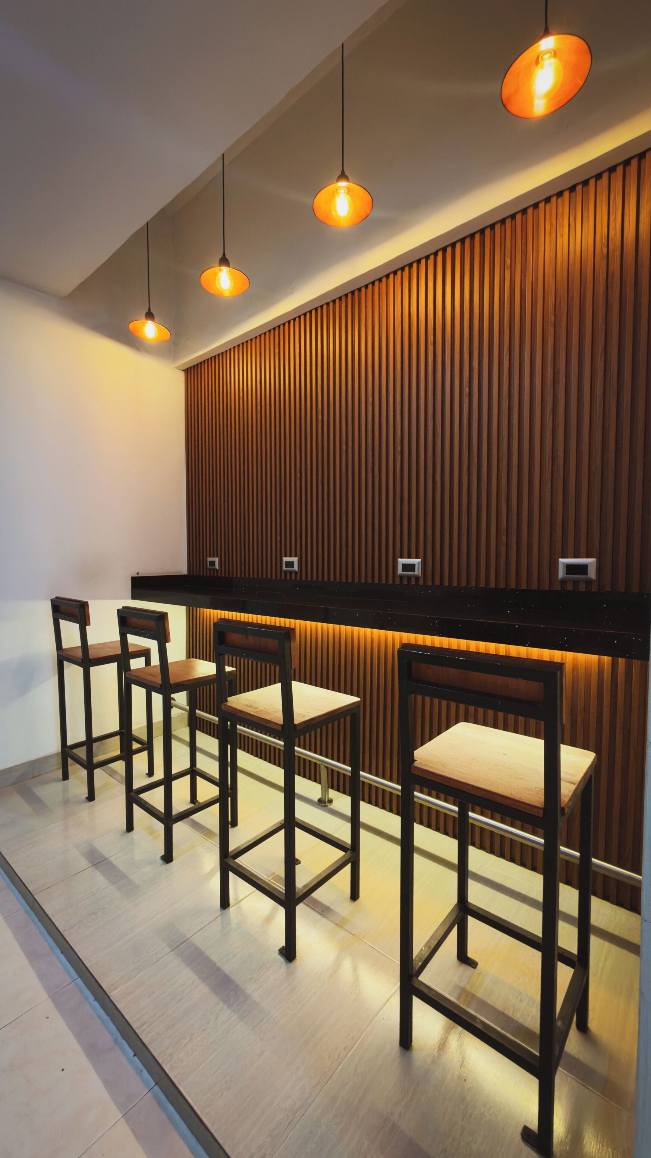 Un espace bar avec un lambris en bois, trois tabourets de bar et des luminaires suspendus.