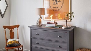 A wooden chair with a patterned cushion, a grey dresser with drawers, a lamp, books, and a framed picture with a crescent moon and the word 'HAW'. - Olive & Stone by Avantstay Private Acre + Hillside Views + Firepit Paso Robles (Templeton)
