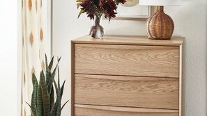 Een houten dressoir met een geweven lamp erop, een vaas met bloemen, en een plant ernaast.