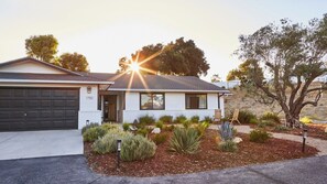 Een gelijkvloers huis met garage, voortuin met diverse beplanting en een boom aan de rechterkant.