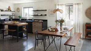 A modern kitchen with a central island, dining table, and chairs. There are shelves above the counter and a window with curtains. - Olive & Stone by Avantstay Private Acre + Hillside Views + Firepit Paso Robles (Templeton)