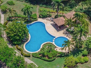A large, irregularly shaped swimming pool surrounded by lush greenery and palm trees, with a small building and lounge area nearby. - Tropical Villa In Hacienda Pinilla (Hacienda Pinilla)
