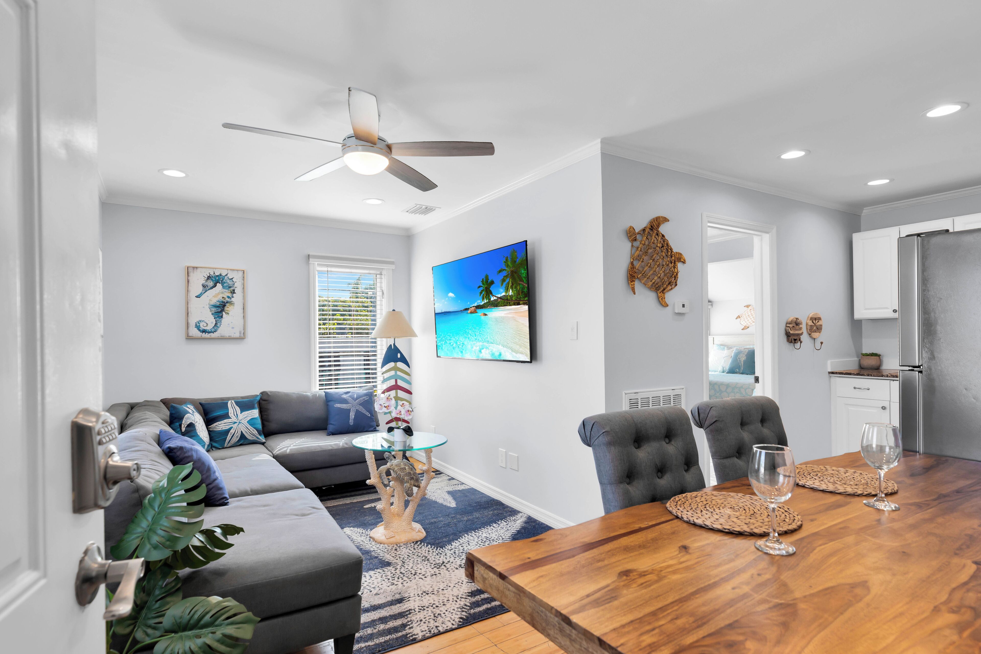 A modern living room with a sectional sofa, a dining table with chairs, a ceiling fan, and a large TV screen displaying a tropical beach scene.