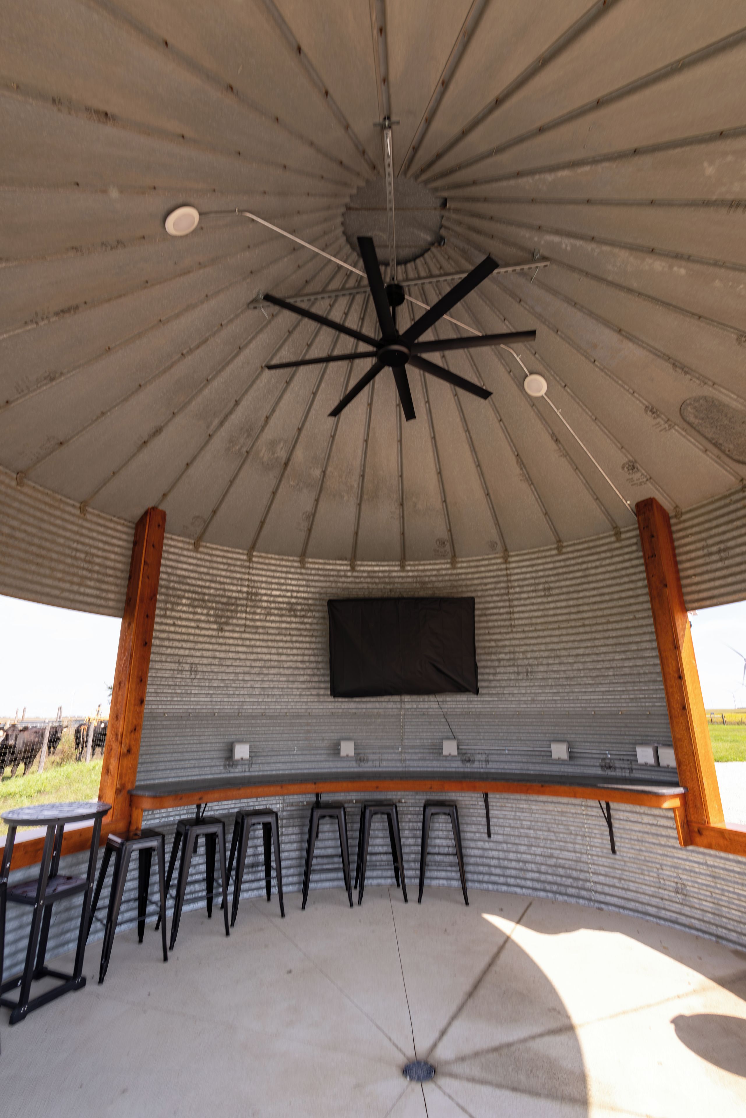 A covered outdoor seating area with a ceiling fan, a mounted television, and a row of black stools.