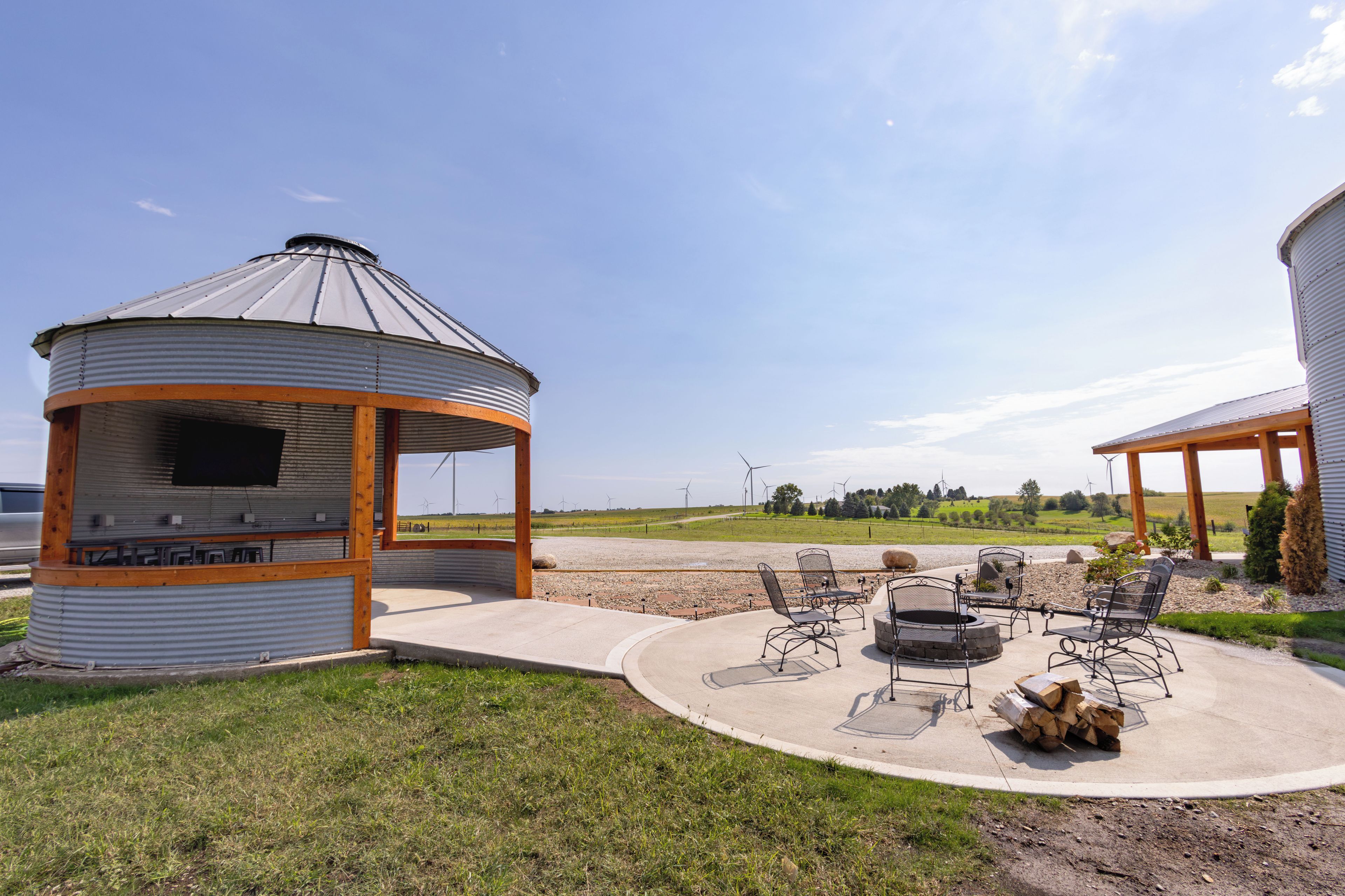 Ein Pavillon mit Grill und Sitzbereich im Freien, umgeben von einer Wiese und Windkraftanlagen im Hintergrund.