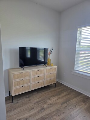 Ein Zimmer mit Flachbildfernseher auf einer hellen Holzkommode, einer Vase mit Trockenblumen und einem Fenster mit Jalousien.