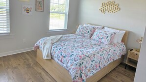 A bedroom with a wooden bed, floral bedding, two windows with blinds, a wall-mounted honeycomb decor, and a bedside table with a lamp. - Bright & Cozy 3BR Near Historic Summerville (Summerville)