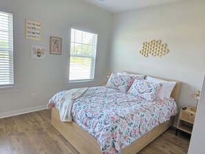Ein Schlafzimmer mit einem hölzernen Bett, floralem Bettzeug, zwei Fenstern mit Jalousien, einer wandmontierten waberartigen Dekoration und einem Nachttisch mit einer Lampe.