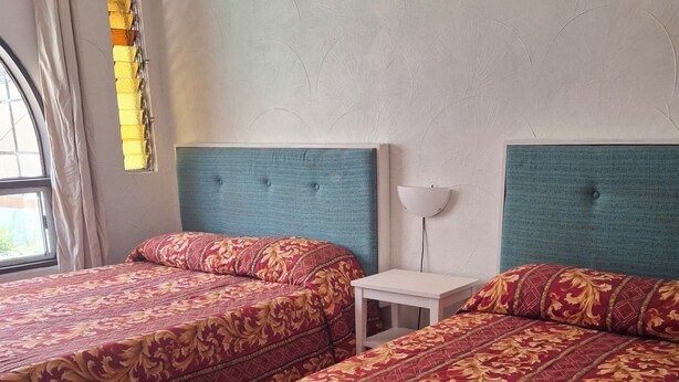 A room with two beds, each with a patterned bedspread, a small bedside table, and a wall-mounted lamp.