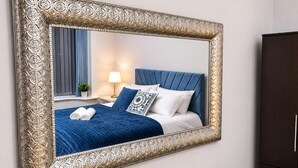 A bedroom with a large ornate mirror, a bed with blue bedding, a nightstand with a lamp, and a dark wardrobe. - Mauldeth Boutique House (Manchester)