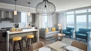Una habitación de hotel moderna con cocina, zona de estar con sofá y sillones, comedor con mesa y sillas, y ventanas grandes que ofrecen vistas.