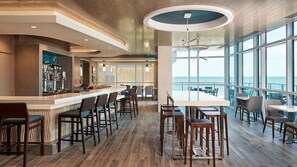 A modern bar area with wooden flooring, bar stools, and a large window overlooking the ocean.