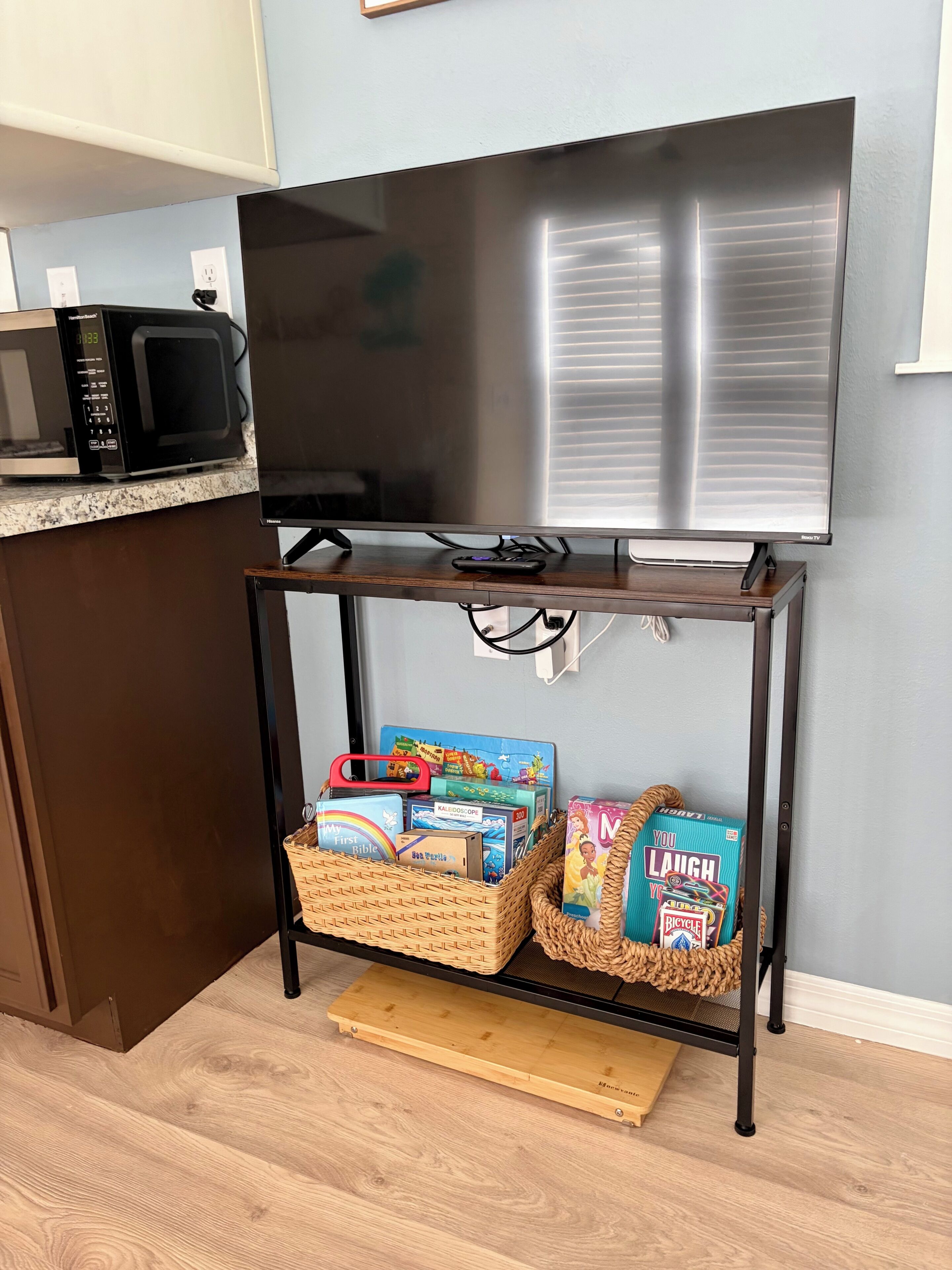 A kitchen with a flat-screen TV, microwave, and a wooden table with baskets containing children's books and toys.
