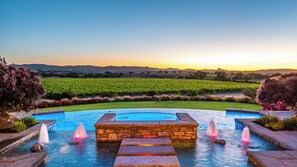 Una piscina con una struttura in pietra al centro, circondata da un giardino curato con vista su un vigneto e delle colline sullo sfondo.