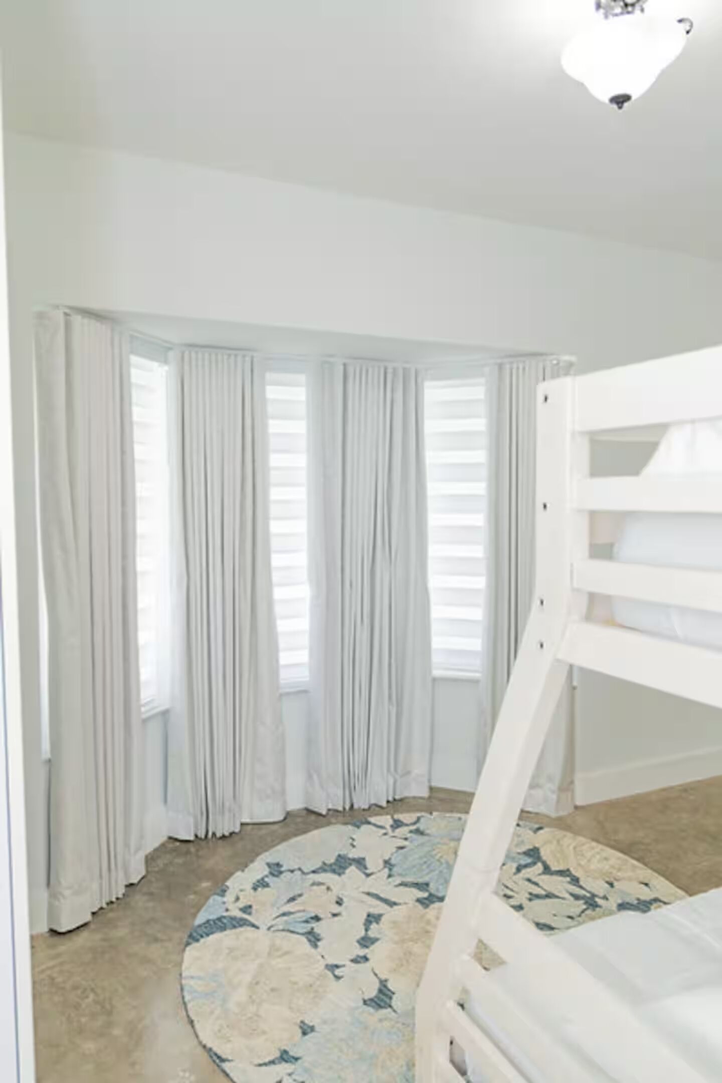 A room with a bunk bed, large windows with curtains, and a patterned rug.