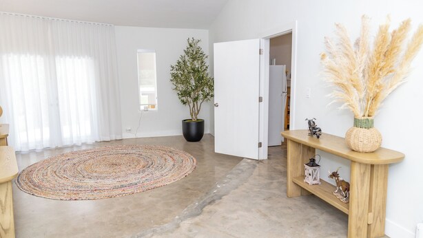 A spacious room with a large window, a potted plant, a round rug, a wooden console table, and a door leading to another room.