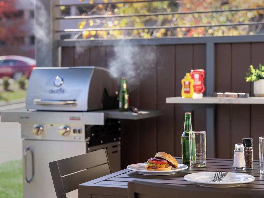 An outdoor grill with a burger on a plate, condiments, and a glass of water on a table.