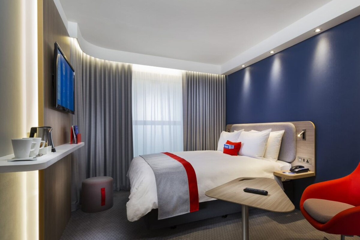 A hotel room with a bed, a red chair, a bedside table with a phone, a TV on the wall, and a window with curtains.