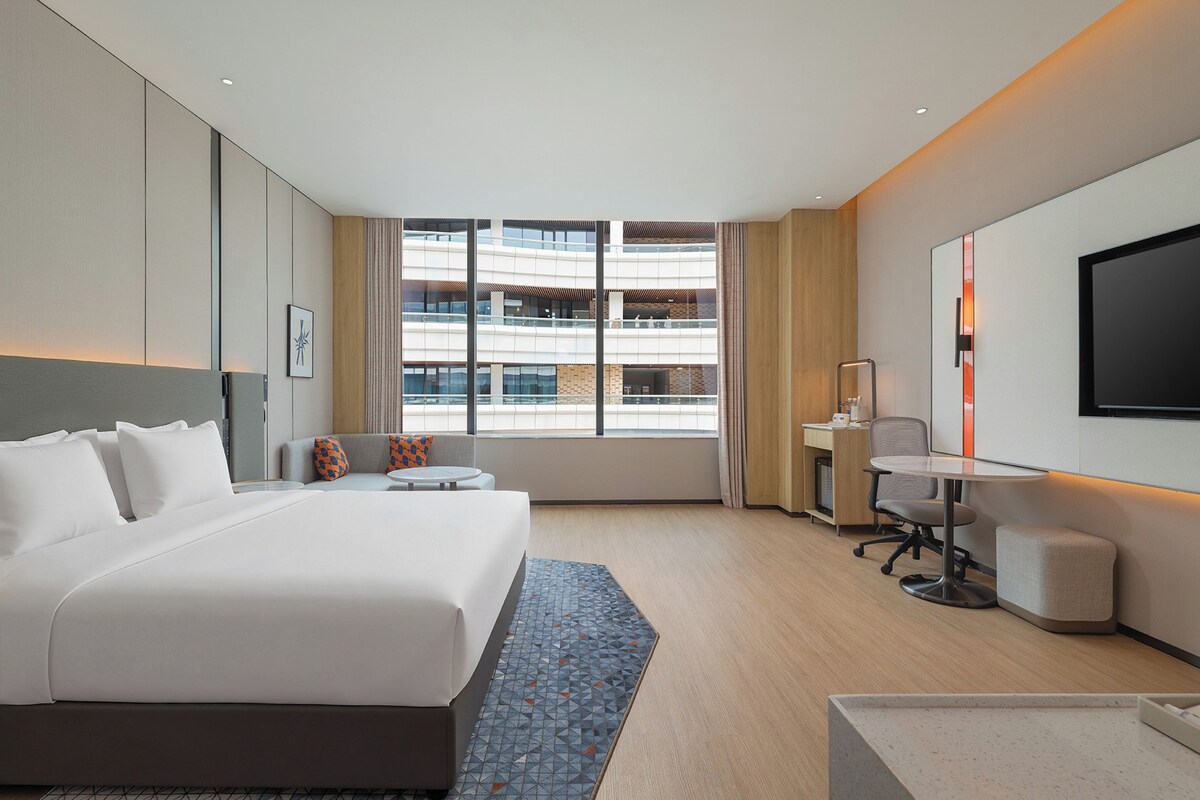 Habitación de hotel con una cama grande, un escritorio, una silla, un televisor y vistas a un edificio de varios pisos.