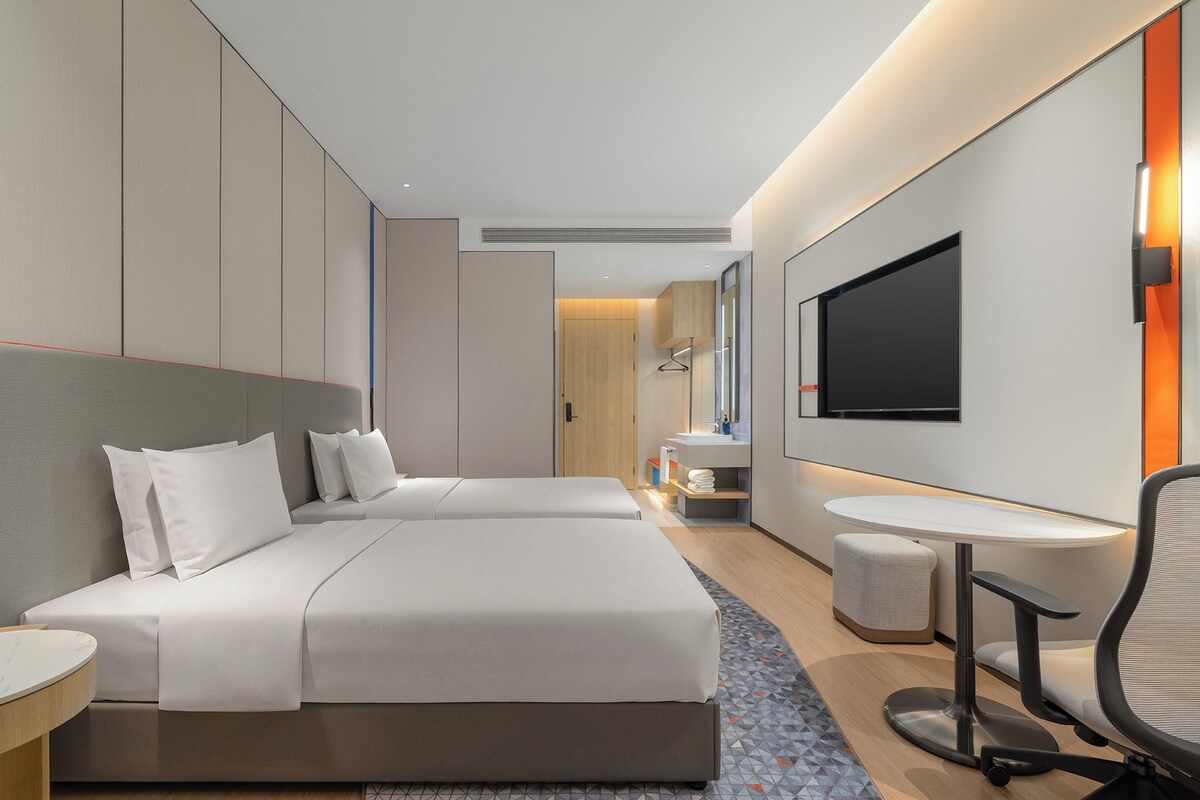 A modern hotel room with a large bed, a small desk, a chair, a TV, and a bathroom visible through a door.