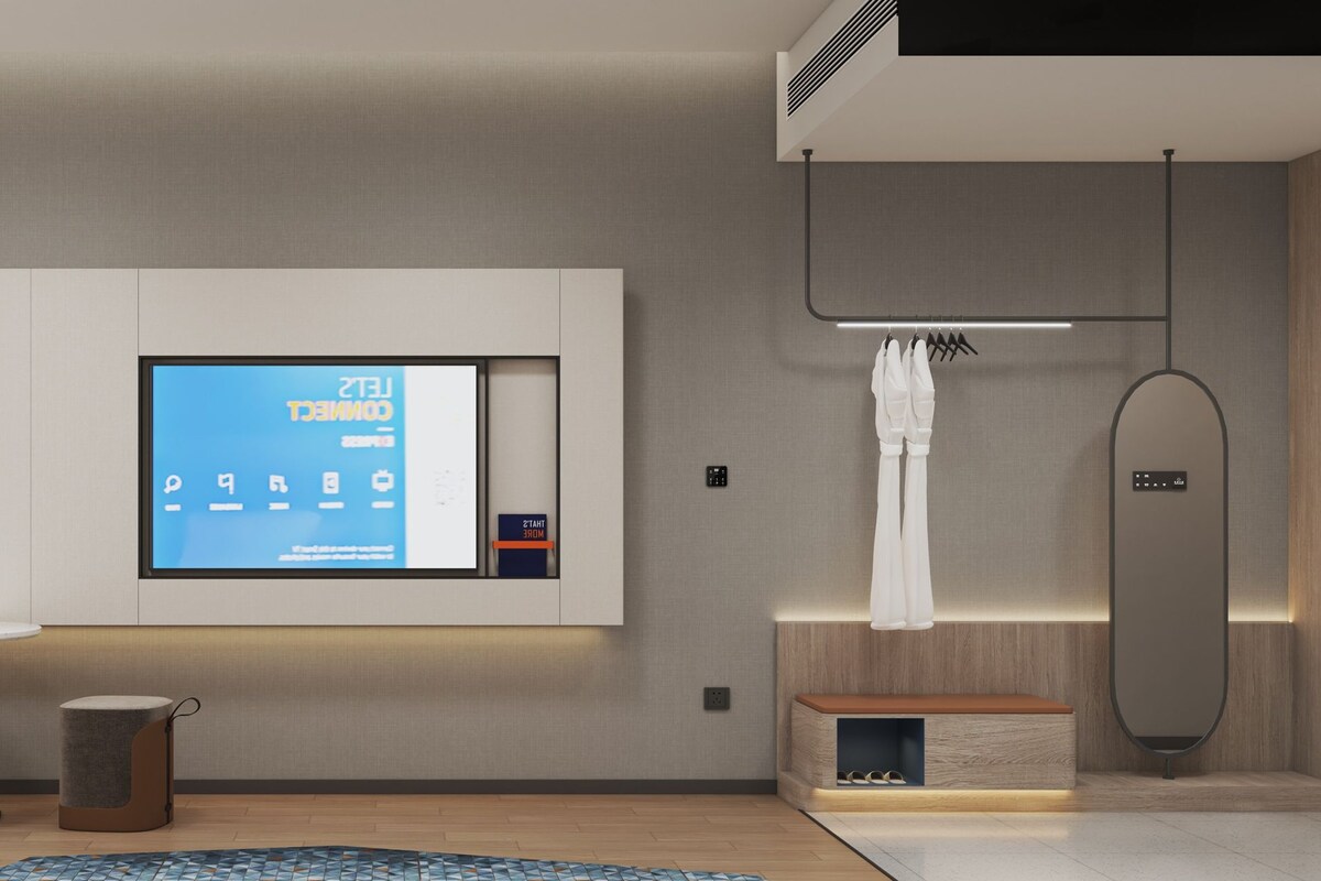A modern hotel room with a large flat-screen TV mounted on the wall, a small round table, a chair, a desk with a coffee machine, a trash bin, a built-in bench, and a shower area with a towel rack.