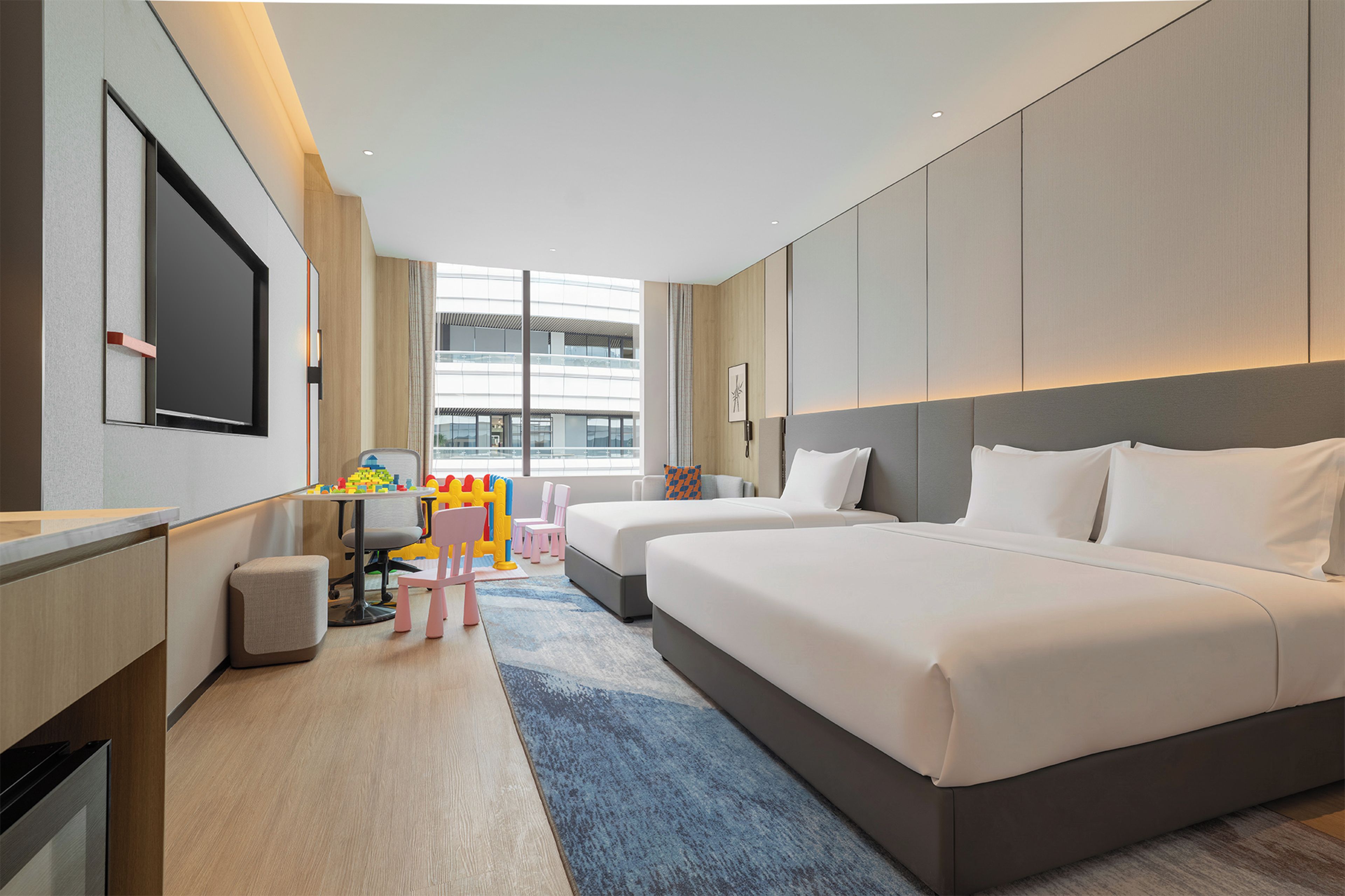 A hotel room with a large bed, a desk with a chair, a television, and a view of the city through large windows.