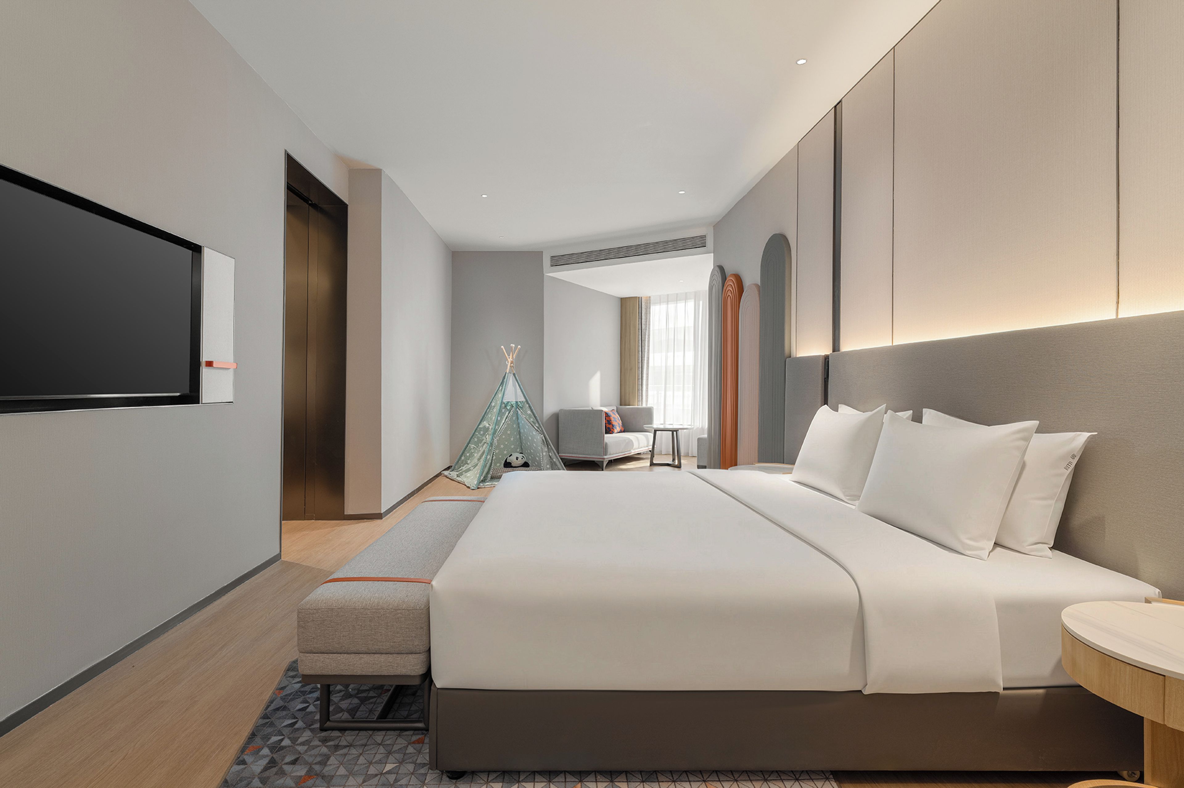 Una habitación de hotel moderna con una cama grande, un televisor de pantalla plana en la pared, un pequeño espacio de descanso con una silla y una mesa, y una estructura decorativa tipo tienda.