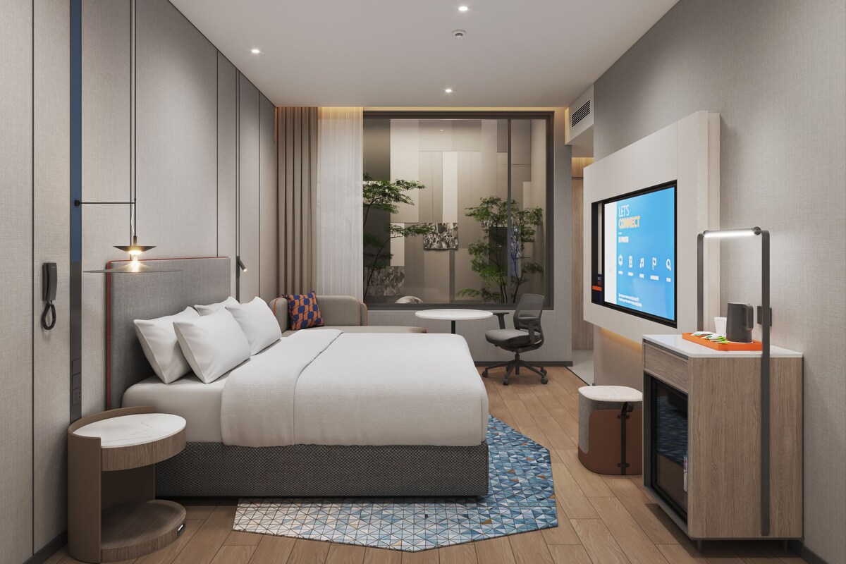 A modern hotel room with a large bed, bedside tables, a TV, and a small seating area.