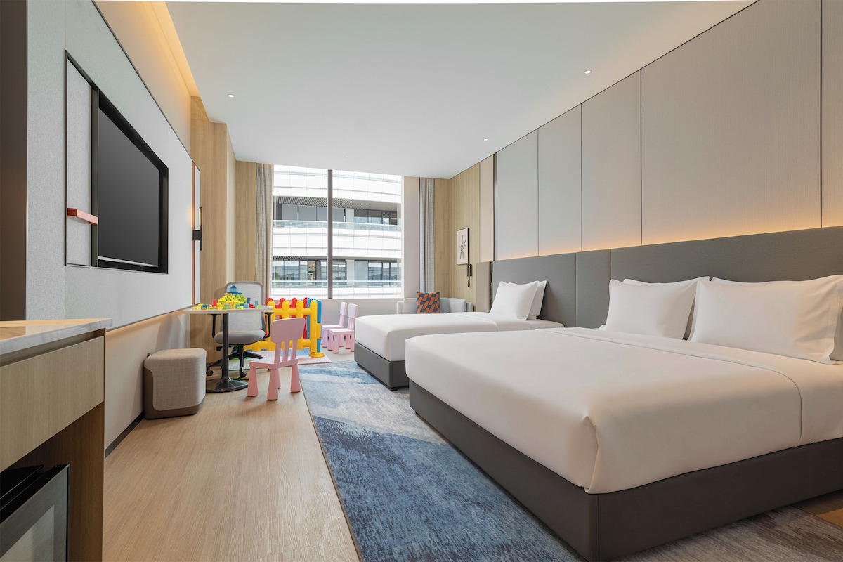 A hotel room with a large bed, a desk with a chair, a television, and a window with a view of buildings.