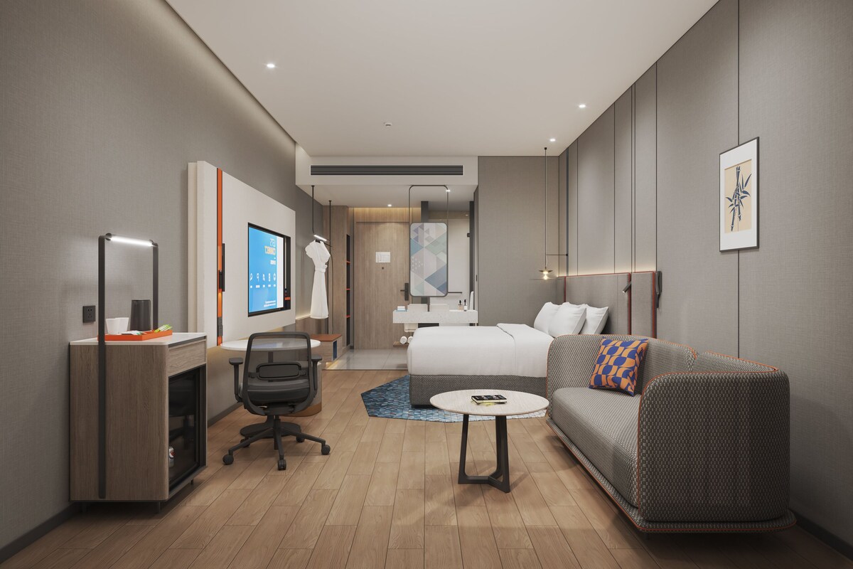 A modern hotel room with a desk, office chair, bed, sofa, and a small round table.