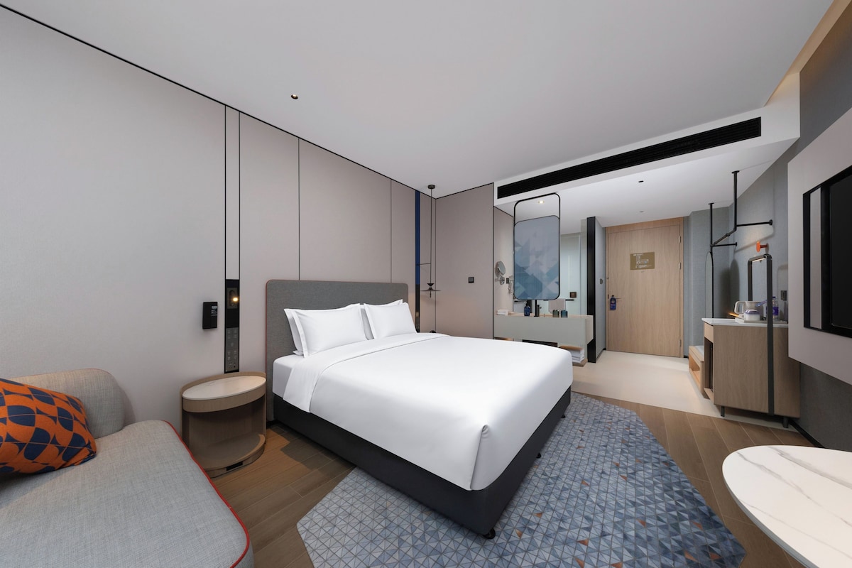 A modern hotel room with a large bed, a bedside table, a bathroom with a mirror and sink, and a separate seating area with a sofa.