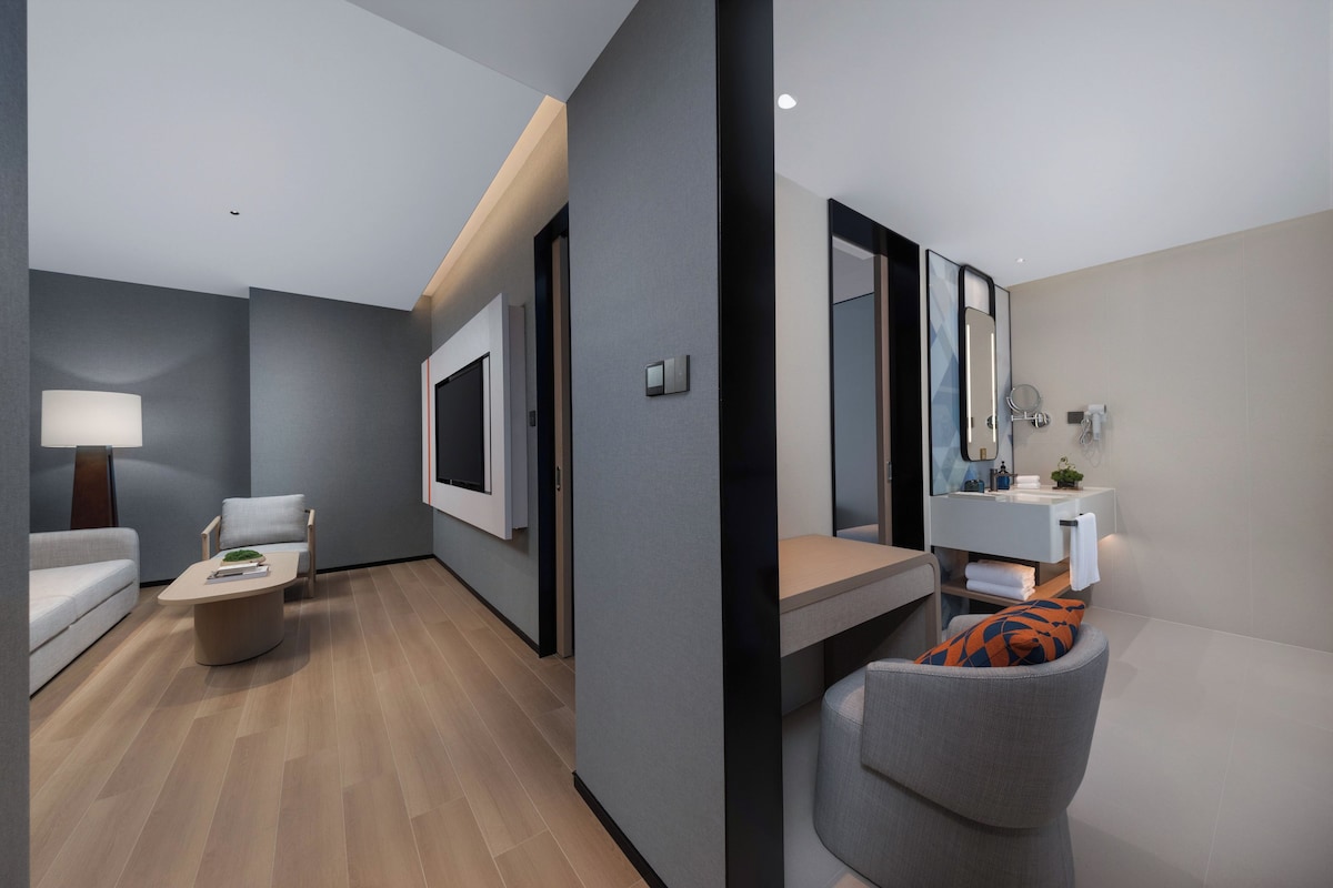 A modern hotel room with a sofa, a small table, a chair, a bed, a bedside table, a mirror, and a bathroom with a sink and towel rack.
