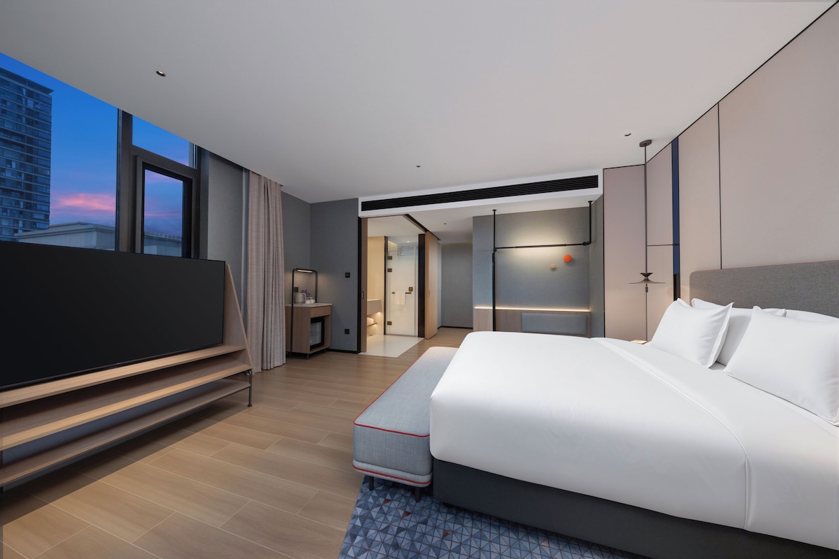 A modern hotel room with a large bed, a flat-screen TV, and a view of the cityscape.