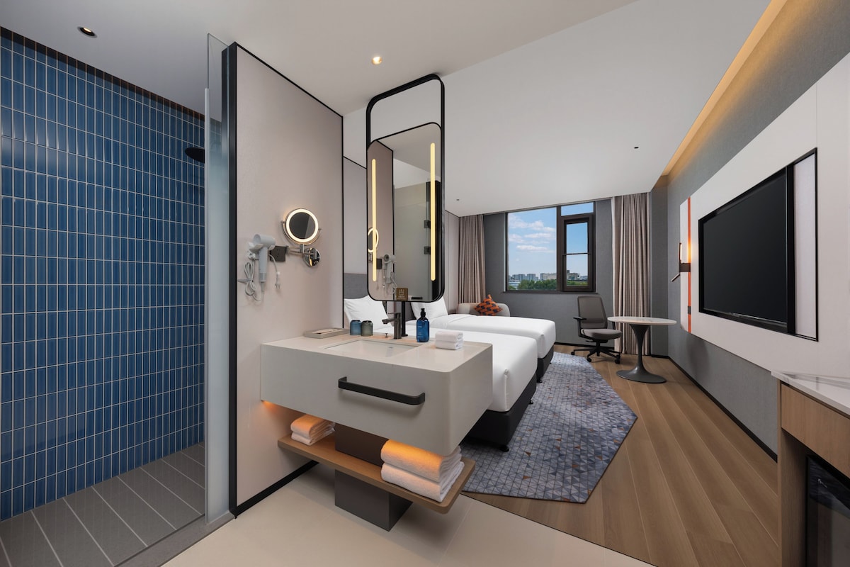 A modern hotel room with a large bed, a separate seating area, a bathroom with a mirror and hairdryer, and a TV mounted on the wall.