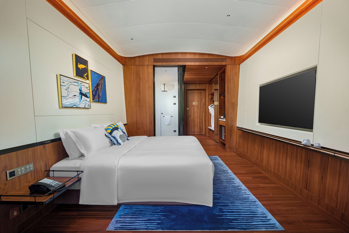 A hotel room with a bed, a desk with a telephone, a blue rug, wooden walls, and artwork on the wall.