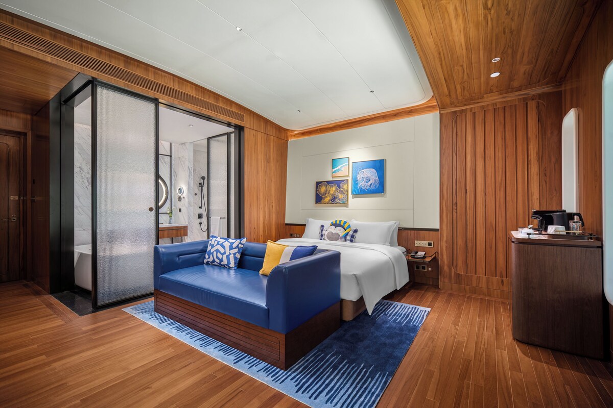 A modern hotel room with a wooden floor, a blue sofa, a bed with white bedding, and artwork on the wall.