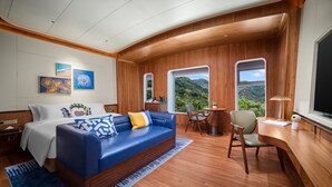 A modern hotel room with a blue sofa, a wooden desk, and a large window offering a view of the outdoors.