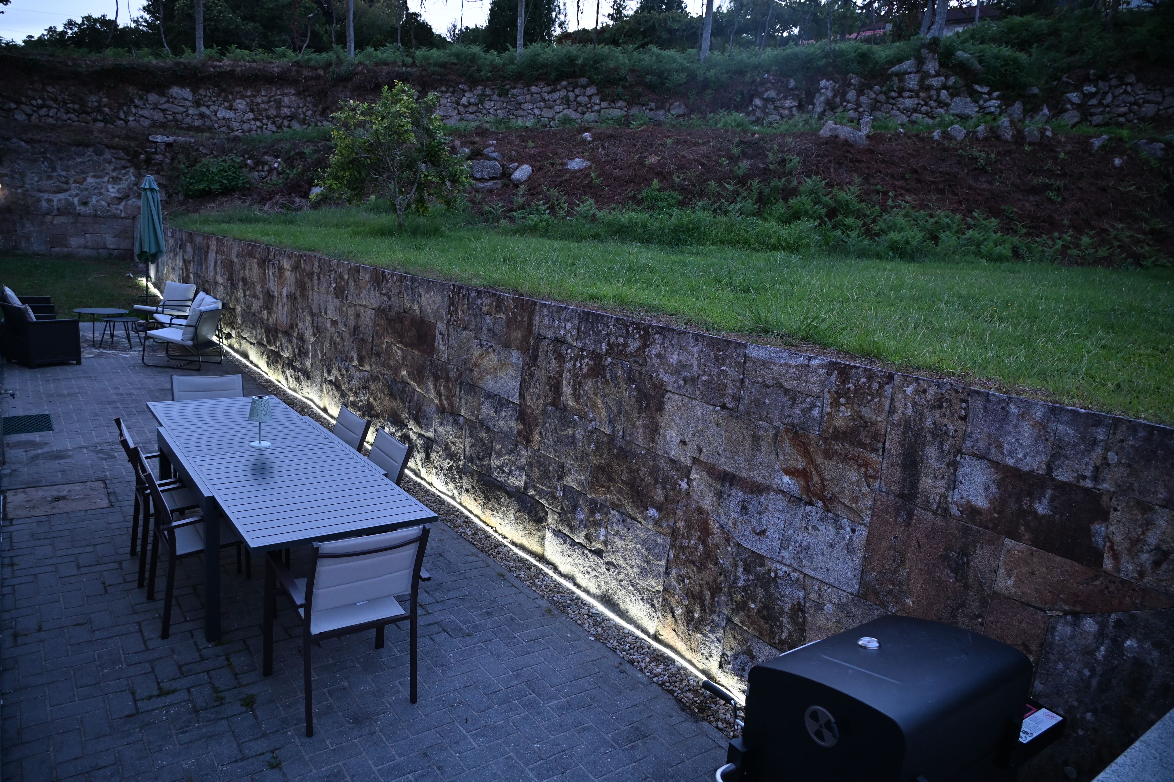 Une terrasse extérieure avec un mur en pierre, une table avec des chaises, un barbecue et un petit parasol.