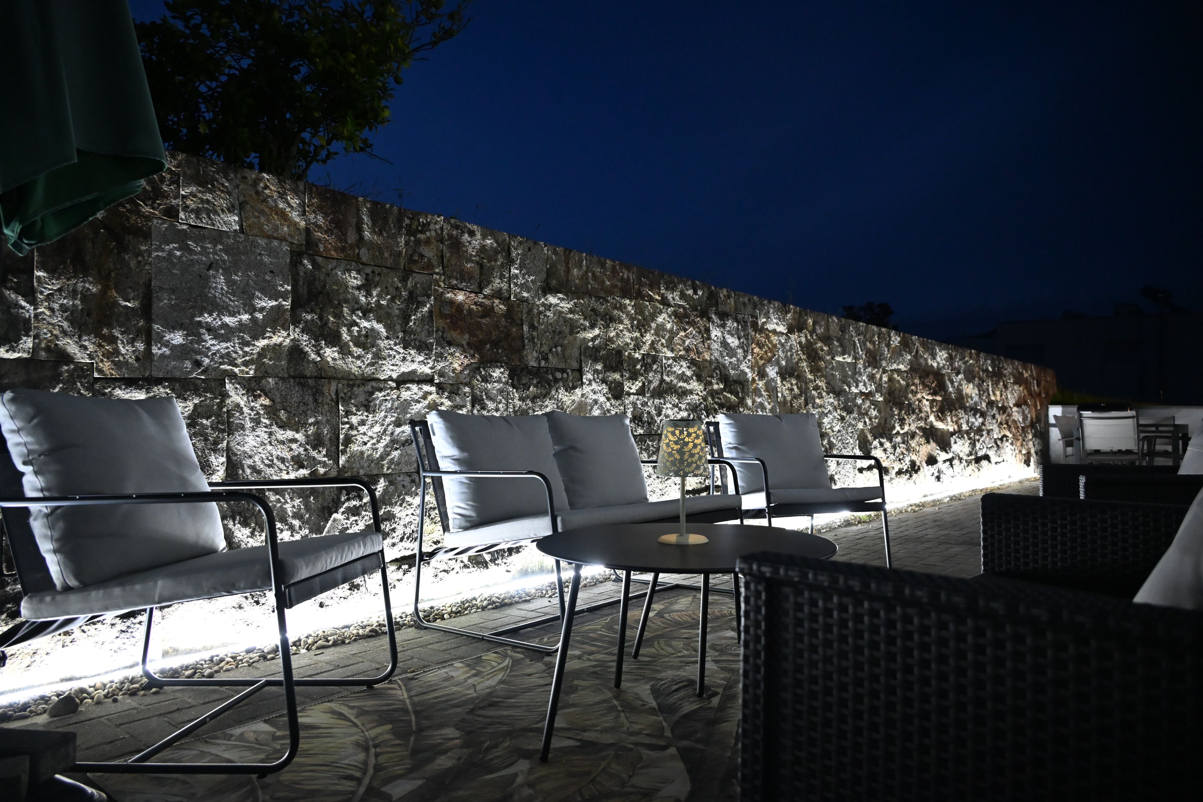 An outdoor seating area with chairs and a table, a stone wall in the background, and a lamp on the table.