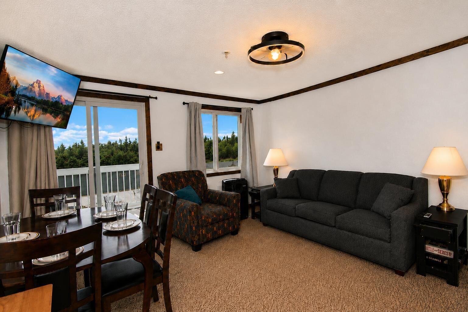 A room with a dining table set for four, a sofa, a TV mounted on the wall, and a window with a view of trees.