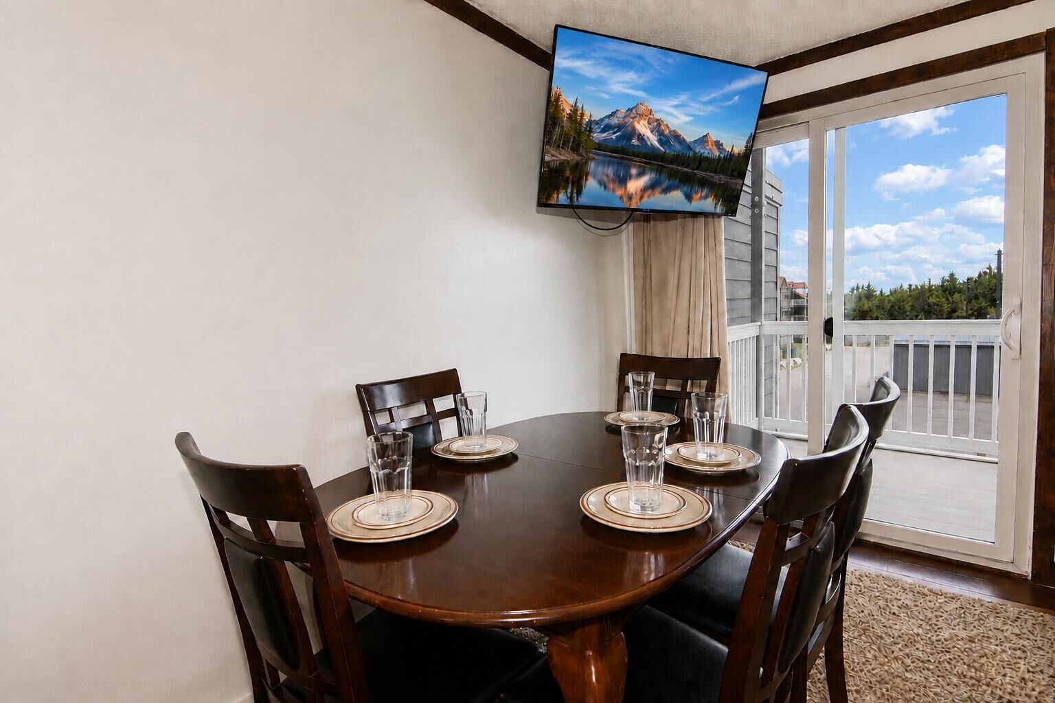 Un coin repas avec une table dressée pour quatre personnes, un téléviseur à écran plat fixé au mur au-dessus, et une vue sur les montagnes par la porte vitrée.