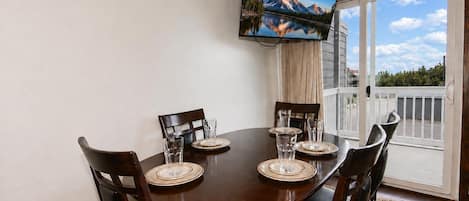 Comedor con mesa dispuesta para cuatro, televisor de pantalla plana sobre ella, y vista de montañas a través de la puerta de vidrio.