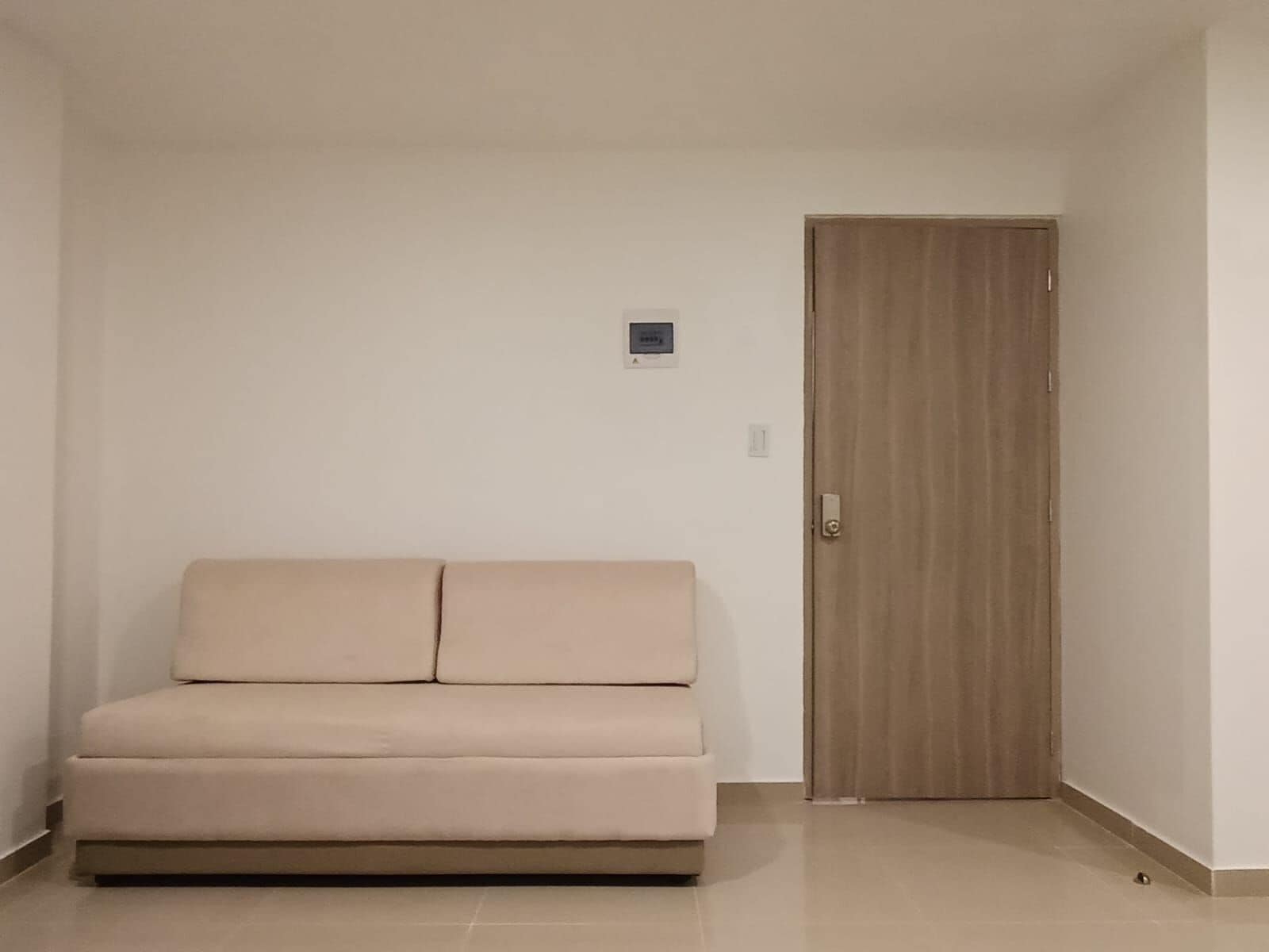 Una habitación en tonos neutros con un sofá color beige, una puerta de madera y un interruptor de luz pequeño, fijado a la pared.