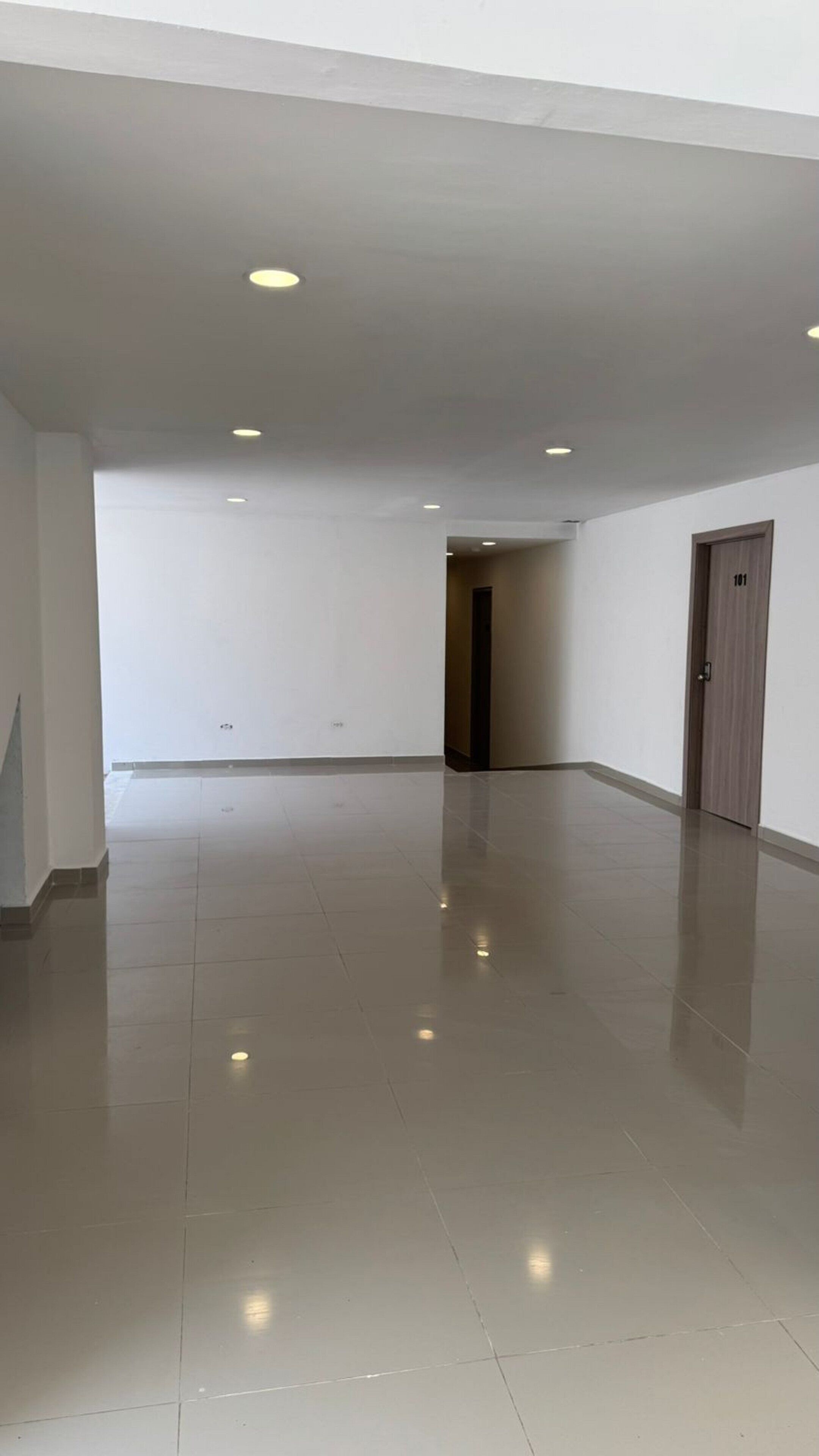 Una habitación amplia y vacía con piso pulido, luces empotradas en el techo y una puerta a la derecha.