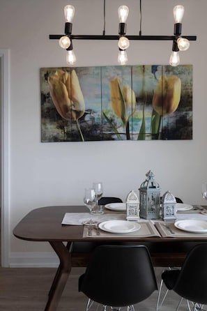 A dining area with a wooden table set for four, a modern light fixture above, and a large floral painting on the wall.