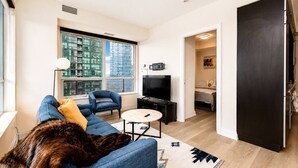 A modern living room with a sofa, a coffee table, a television, and a large window with a city view. - Cozy 2BR 2BA Condo | Steps to Subway | Pool & Gym (Toronto)