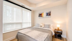 A bedroom with a bed, a nightstand, a lamp, a window with blinds, and a painting on the wall. - Cozy 2BR 2BA Condo | Steps to Subway | Pool & Gym (Toronto)