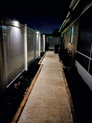 A pathway leading to a building with a covered area, possibly a patio or deck, and a trash bin.